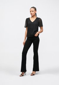 Donna in piedi con la mano in tasca che indossa una maglia nera trasparente a maniche corte, pantaloni neri aderenti e sandali neri con tacco alto su sfondo bianco.