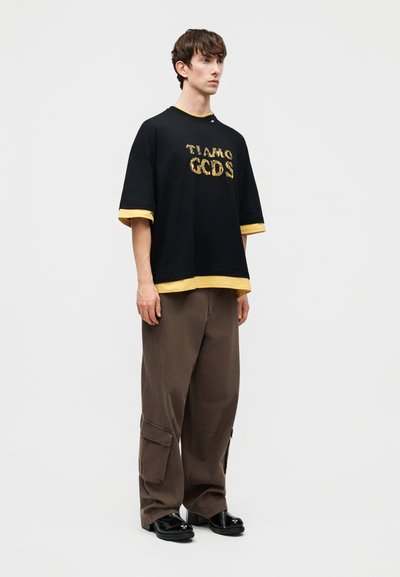 Junger Mann steht und trägt ein schwarz-gelbes T-Shirt mit mehreren Lagen und dem Schriftzug "GCDS" sowie weite braune Cargohosen und schwarze Schuhe.