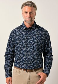 Mann mit grauem Haar und Bart, der ein langärmeliges, marineblaues, floral gemustertes Hemd und beige Hosen mit einem braunen Gürtel trägt, vor einem einfarbigen Hintergrund.