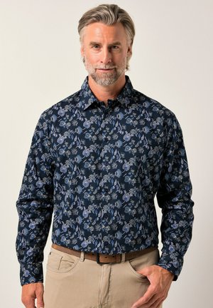 Homme aux cheveux et à la barbe grises portant une chemise à manches longues à motifs floraux bleu marine et un pantalon beige avec une ceinture marron, sur fond uni.