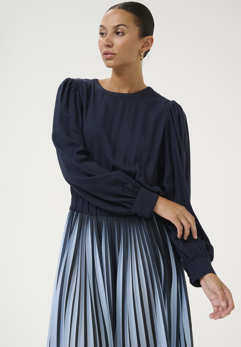 Tumesinane pikkade varrukatega särk, millel on plisseeritud õlavarred ja tihe vöökoht, koos plisseeritud seelikuga, millel on gradient sinised triibud.