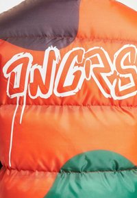 Farverig polstret jakke med en orange gradient og store grønne og lilla former samt hvid graffiti-stil tekst. Glat overflade.
