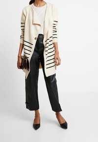 Jasno beżowy kardigan z czarnymi paskami, rozpinany z przodu, z jednym guzikiem. Połączony z spodniami z wysokim stanem z czarnej skóry i czarnymi szpilkami w szpic.