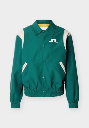 Μπουφάν bomber σε τυρκουάζ πράσινο με μπεζ λεπτομέρειες στους ώμους και τις τσέπες, κλείσιμο με κουμπιά τύπου snap, ελαστικά μανσέτες και τελείωμα, και λευκό λογότυπο στο στήθος.