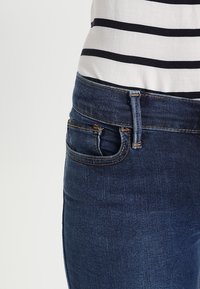 Acercamiento de unos pantalones vaqueros oscuros con una camisa a rayas blanca y azul marino parcialmente visible, destacando los detalles de la cintura y el bolsillo.