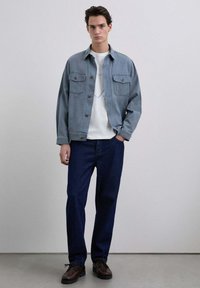 Chaqueta de mezclilla azul claro con dos bolsillos en el pecho, usada sobre una camiseta blanca con un gráfico lineal azul, combinada con pantalones azul oscuro y zapatos marrones.