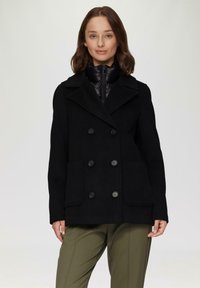 Schwarzer Wollmischung-Peacoat mit doppelreihigen Knöpfen, breitem Revers und zwei Fronttaschen, getragen über einer schwarzen gepolsterten Innenjacke.