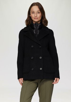 Manteau en laine noir mélangé avec des boutons double boutonnage, larges revers et deux poches avant, porté par-dessus une veste intérieure rembourrée noire.
