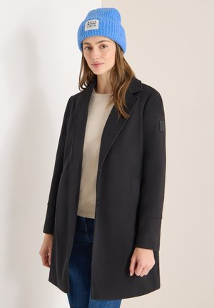 Cappotto nero con design sartoriale, caratterizzato da colletto a revers, tasche laterali e un berretto blu lavorato a maglia con etichetta.