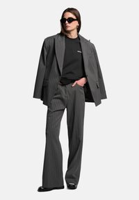 Šedý pruhovaný oblek s širokými kalhotami a oversized sako, spárovaný s černým tričkem a elegantními slunečními brýlemi. Hladký textilní povrch.