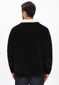 Fekete kordbársony dzseki krém színű műszőrme gallérral, egyenes szabással és látható varrási részletekkel, sötét farmernadrág felett viselve.