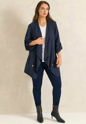 Femme portant un cardigan navy drapé à ouverture frontale sur un haut blanc, un jean skinny navy et des bottines noires à talons.