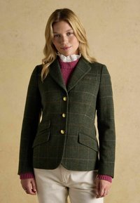 Vihreä villablazeri, jossa on ruudullinen kuvio, kultanapit ja kaksi etutaskua. Päällä vaaleanpunainen neulepaita, jossa on frillavalkoinen kaulus.