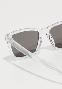 Lunettes de soleil en plastique transparent avec des verres gris foncé, verres triangulaires et branches droites transparentes. Les accents incluent de petits détails au niveau des charnières.