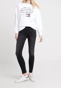 Felpa bianca con testo nero e un piccolo logo a strisce, abbinata a jeans skinny neri e sneakers bianche con un accento rosso.