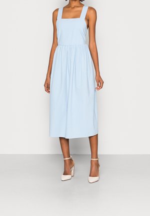 Mujer que lleva un vestido midi azul claro sin mangas con tirantes anchos y zapatos beige de tacón alto con pulsera en el tobillo, de pie frente a un fondo liso.