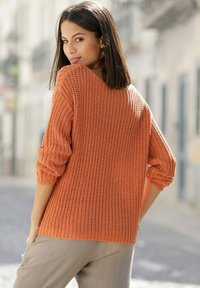 Oranger gestrickter Pullover mit langen Ärmeln, strukturiertem Muster und lockerer Passform. Stylisch kombiniert mit hellen Hosen, von hinten betrachtet.
