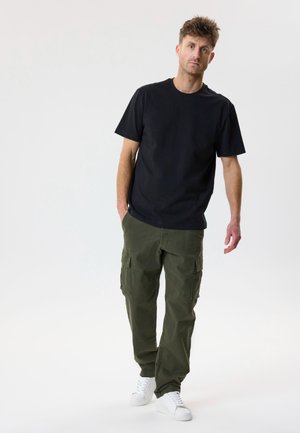 Mann trägt schwarzes T-Shirt, grüne Cargo-Hose und weiße Turnschuhe, steht mit einer Hand in der Tasche vor einfarbigem Hintergrund.