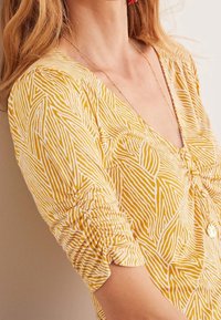 Žlutá blůza s geometrickým bílým vzorem, výstřihem do V a nařasenými detaily na rukávech. Lehká tkanina s hladkým povrchem.