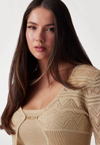 Beige knitted sweater with a scoop neckline and textured patterns, featuring a decorative pin with the brand name positioned at the bust.