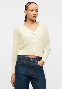 Jeune femme aux cheveux tressés portant un cardigan court en tricot crème avec trois boutons et un jean taille haute bleu marine, la main dans la poche.