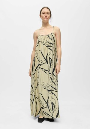 Femme portant une robe longue beige avec des motifs abstraits de feuilles noires, fines bretelles, debout devant un fond blanc uni.