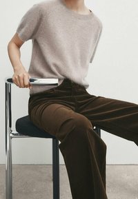 Pull en cachemire beige à manches courtes, associé à un pantalon en velours côtelé large marron foncé. Modèle assis sur une chaise en métal.