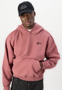 Hoodie rose poussiéreux avec une poche avant, logo noir et accents cloutés sur les manches. Associé à un pantalon noir avec un motif craquelé.