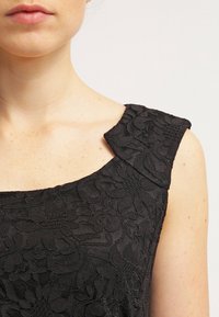 Vestido negro de encaje con un patrón floral texturizado, que presenta un escote redondeado y un diseño de hombros anchos y estructurados.