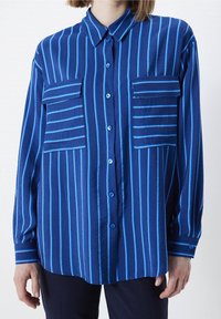 Donna che indossa una camicia button-up a maniche lunghe blu con strisce verticali azzurre e due grandi tasche sul petto con alette.