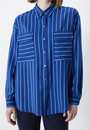 Donna che indossa una camicia button-up a maniche lunghe blu con strisce verticali azzurre e due grandi tasche sul petto con alette.