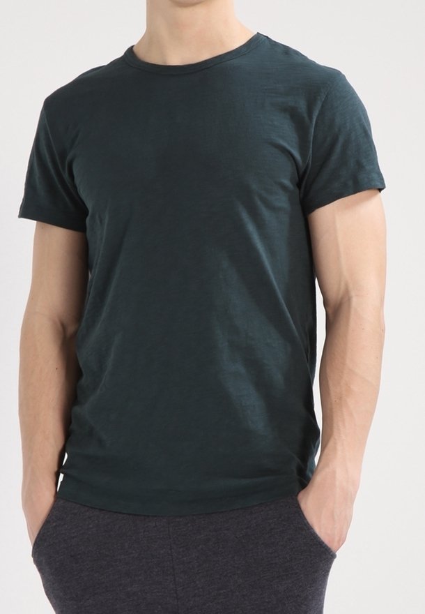 Homme portant un t-shirt uni vert foncé à manches courtes et un pantalon gris foncé, les mains dans les poches, debout devant un fond clair uni.
