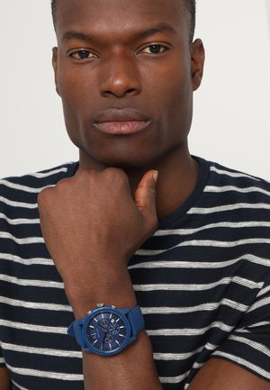 Mężczyzna z założonymi ramionami, ubrany w niebiesko-białą koszulę w paski oraz niebieski zegarek chronograf z silikonowym paskiem.