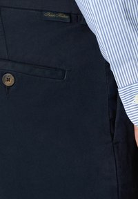 Granatowe spodnie o teksturowanym wykończeniu, z zapięciem na guzik i małą metką Brooks Brothers na pasku. Widoczny rękaw w paski.