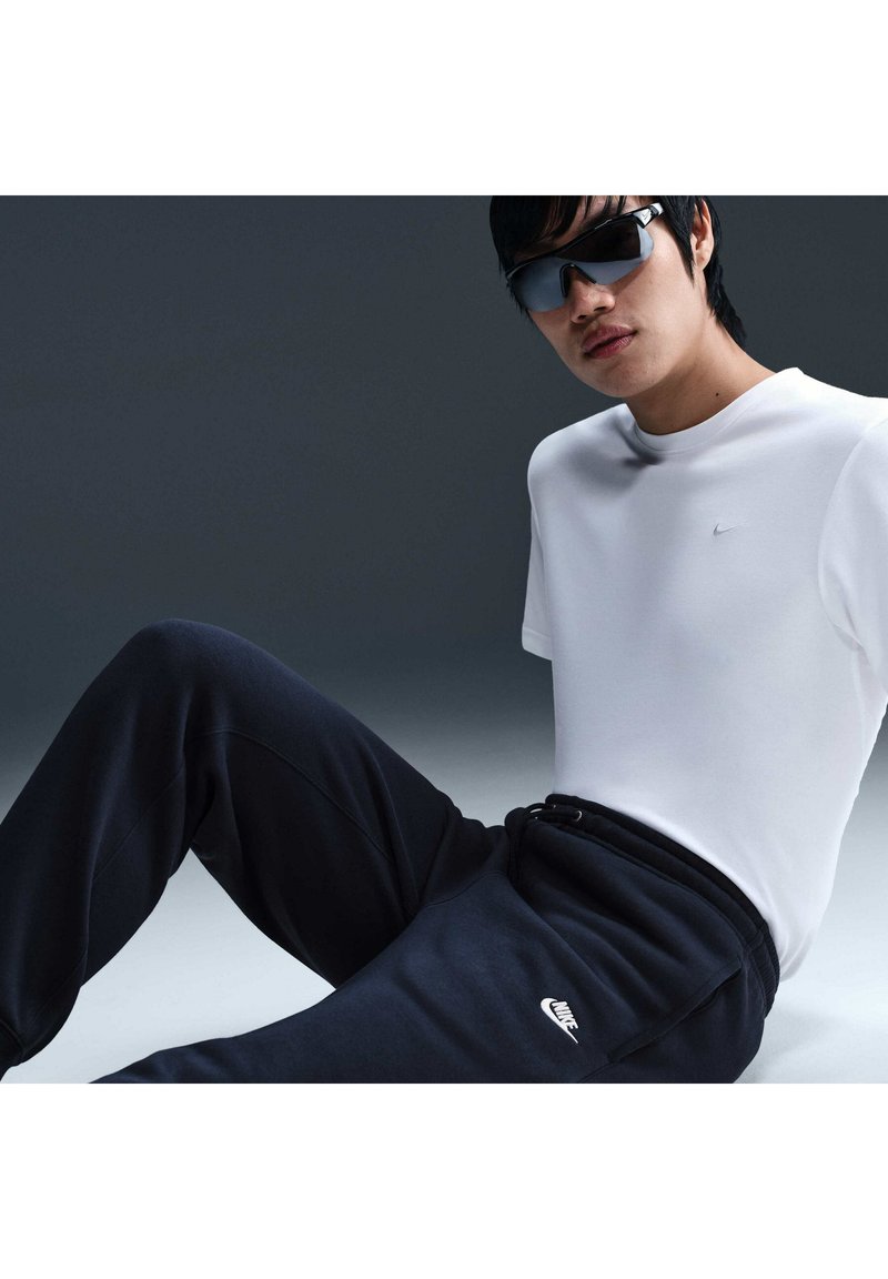 Persona che indossa occhiali da sole neri, una t-shirt bianca e pantaloni della tuta neri Nike è seduta contro uno sfondo scuro, guardando verso la fotocamera.