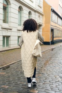Gilet long matelassé beige, haut à manches longues gris, pantalon noir et baskets blanches. Portant un tote bag en toile léger. Rue pavée.