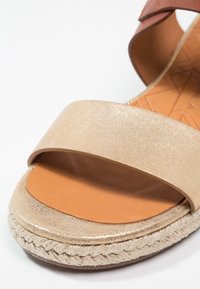 Sandale à bout ouvert avec une bride métallique dorée et une bride de cheville marron, avec une semelle en jute tressé et une semelle intérieure en cuir lisse.