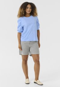 Világoskék puffujjú ing fekete-fehér csíkozott sorttal párosítva. A modell világos bézs mokaszinokat visel. Minimális háttér, egyenletes világítással.