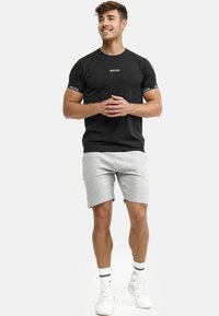 Sort bomulds t-shirt med logo på brystet og mønstrede ærmer, parret med lysegrå shorts. Modellen bærer hvide sko med stribede sokker.