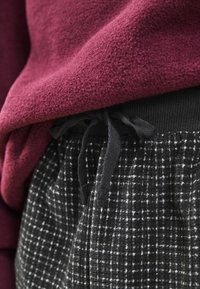 Burgundin värinen fleece-paita mustien ruudullisten housujen päällä, joissa on musta joustovyö vyötäröllä. Pehmeä rakenne ja löysä istuvuus ovat nähtävissä.