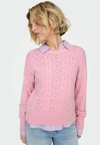 Pull rose en maille avec motifs de cœurs, ourlet et poignets côtelés. Porté sur une chemise à col rayée bleu clair et blanc. Look décontracté.