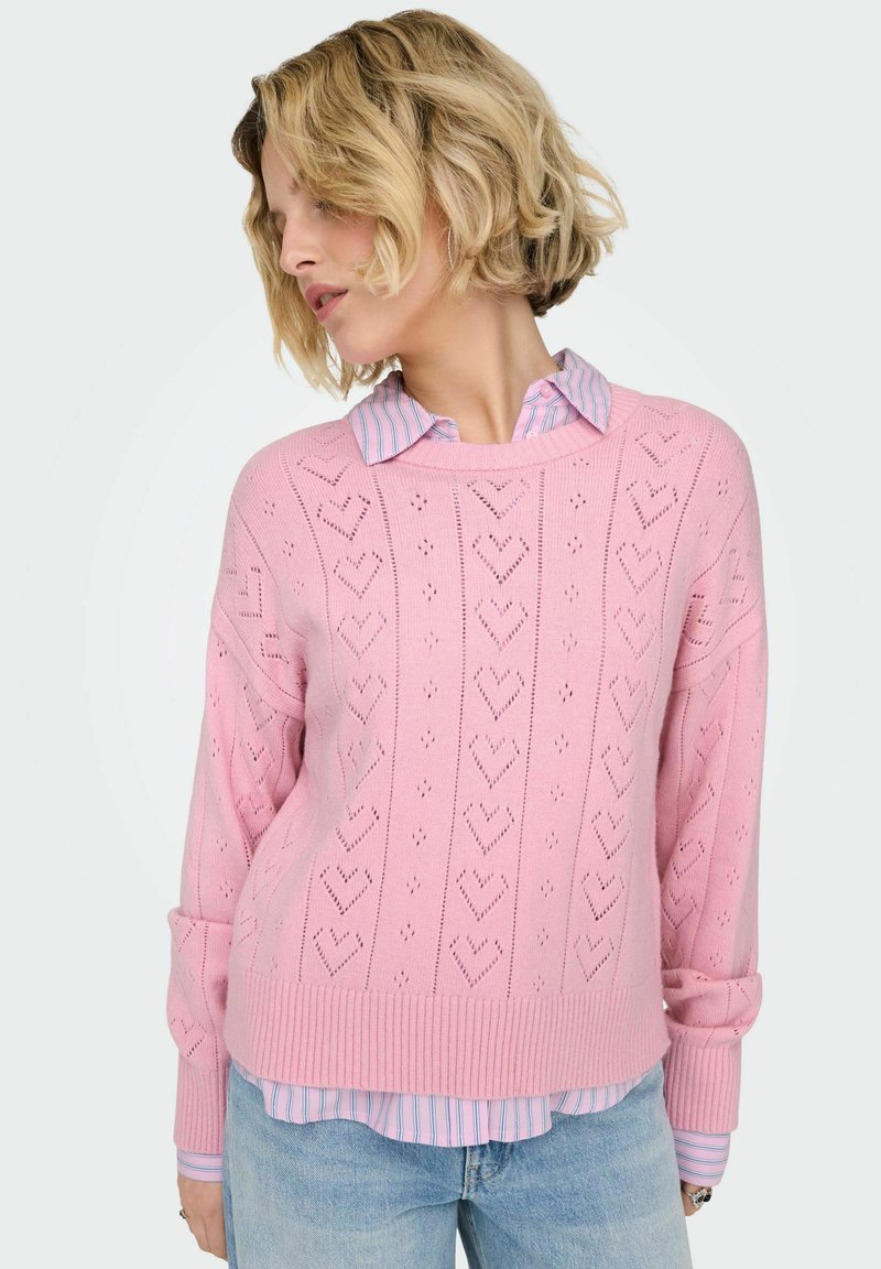 Pull rose en maille avec motifs de cœurs, ourlet et poignets côtelés. Porté sur une chemise à col rayée bleu clair et blanc. Look décontracté.