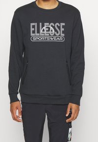 Sweat-shirt gris foncé à encolure ronde, avec un logo blanc "ELLESSE SPORTSWEAR" et des poches latérales zippées. Tissu doux, poignets côtelés.