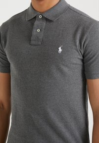 Grå polotröja med mjuk textur, med klassisk krage, två knappar och en vit broderad logotyp på vänster bröst.