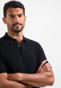 Zwarte polo met korte mouwen, twee knopen bij de kraag, een zachte textuur en een slim fit, gedragen door een model met gekruiste armen.