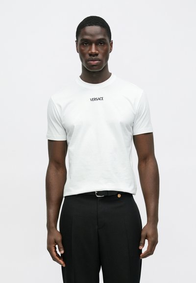 Biały bawełniany t-shirt z krótkimi rękawami, z czarnym logo "VERSACE" umieszczonym centralnie nad klatką piersiową. W połączeniu z czarnymi, dopasowanymi spodniami.