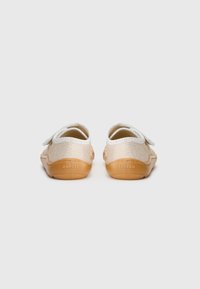 Zapatos infantiles con un upper textil en beige claro y dorado, cierre con tira elástica, punta redondeada y suela de goma marrón. Diseño minimalista.
