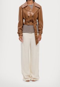 Bruine leren dubbelgeknoopte trenchcoat met een ceintuur in de taille, gecombineerd met een gestreept topje en brede crèmekleurige broek. Gladde textuur en een nauwsluitende pasvorm.