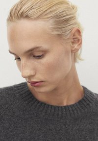 Pull en laine gris avec un col côtelé. Le sujet a des cheveux blonds clairs et des taches de rousseur visibles sur un teint lisse, regardant vers le bas.