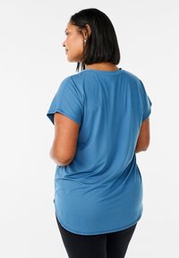 Blauwe short-sleeve shirt van zachte stof, met een relaxte pasvorm, een ronde onderkant en zijsplitten voor gemak in beweging, gedragen over zwarte broeken.